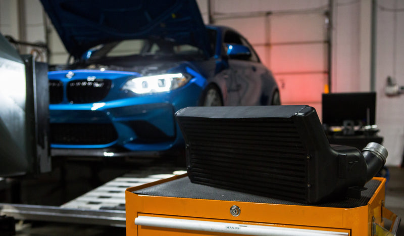 Mishimoto BMW front mount intercooler core on a shop cart with a BMW on the dyno behind it